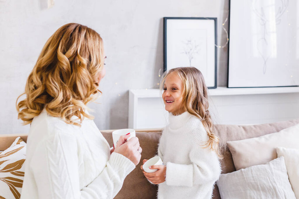 Mother and daughter in light sweaters are sitting on the sofa, the room is decorated for the new year and Christmas, hugs, conversations, smiles, kisses, waiting for a gift and magic - Zdjęcie, obraz