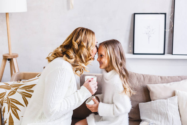 Mother and daughter in light sweaters are sitting on the sofa, the room is decorated for the new year and Christmas, hugs, conversations, smiles, kisses, waiting for a gift and magic - Photo, image
