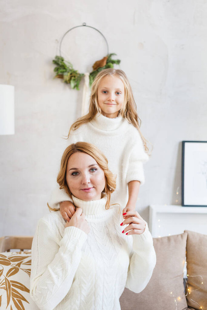 Mother and daughter in light sweaters are sitting on the sofa, the room is decorated for the new year and Christmas, hugs, conversations, smiles, kisses, waiting for a gift and magic - Photo, Image