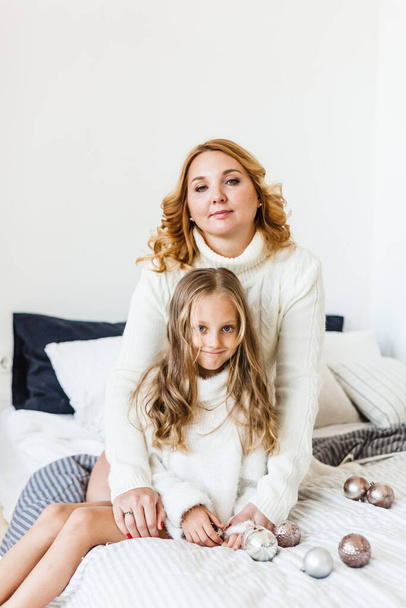 Mom and daughter blonde hair and white sweaters lie on the bed, light up, say toys for Christmas and new year tree, Christmas tree decoration - Photo, image
