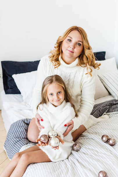 Moeder en dochter blond haar en witte truien liggen op het bed, licht op, zeggen speelgoed voor Kerstmis en nieuwjaar boom, kerstboom decoratie - Foto, afbeelding