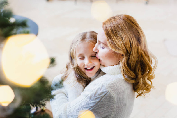 Een meisje met haar moeder in de buurt van de kerstboom, het interieur ingericht voor het nieuwe jaar en Kerstmis, familie en vreugde, tradities - Foto, afbeelding