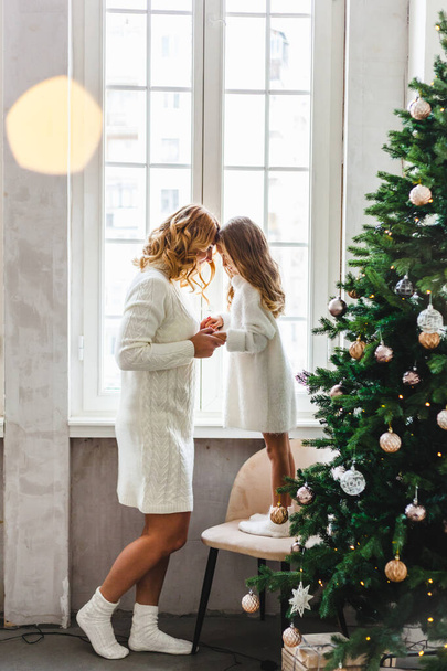 Girl child and mother are standing near the window, hugging, waiting for guests or Santa Claus for new year, Christmas, the room is decorated with a Christmas tree and balloons - Photo, Image
