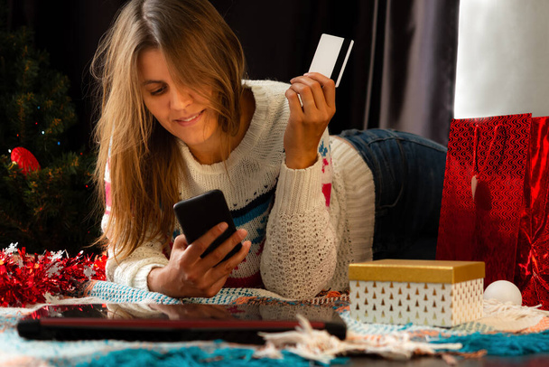 Μια μελαχρινή κοπέλα με χειμωνιάτικο πουλόβερ χαμογελάει στο σπίτι και κοιτάζει το κινητό της. Χριστουγεννιάτικα online ψώνια. Αγοράστε online δώρα για το νέο έτος. το εσωτερικό του σπιτιού με μια ευτυχισμένη γυναίκα. - Φωτογραφία, εικόνα