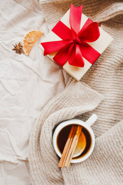 затишна різдвяна листівка гігієни. порцелянова чашка чаю з апельсиновим шматочком, паличками кориці, зіркою анісу на бежевому покритті, подарунком з червоною стрічкою та блискучою гірляндою на фоні, вибірковий фокус
 - Фото, зображення