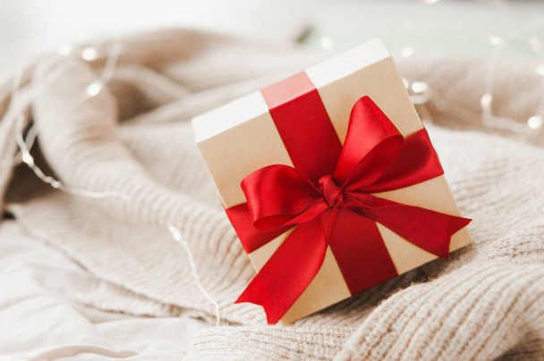 stylish square beige box with red festive satin ribbon on beige crumpled blanket and sparkling garland on background, selective focus - Foto, imagen