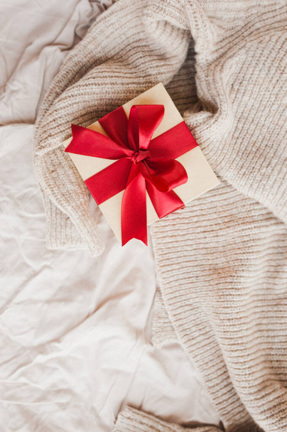 stylowe kwadratowe beżowe pudełko z czerwoną świąteczną satynową wstążką na beżowym, przytulnym, zgniecionym kocu selektywna ostrość - Zdjęcie, obraz