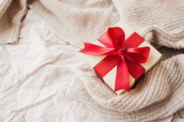 elegante caja beige cuadrada con cinta de satén festivo rojo en color beige acogedor manta arrugada enfoque selectivo - Foto, imagen