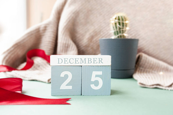 wooden calendar cubes with date December 25, red ribben on beige cozy sweater and small cute green cactus decorated with garland in pot. cozy simple desktop decor at Christmas time, selective focus - Foto, afbeelding