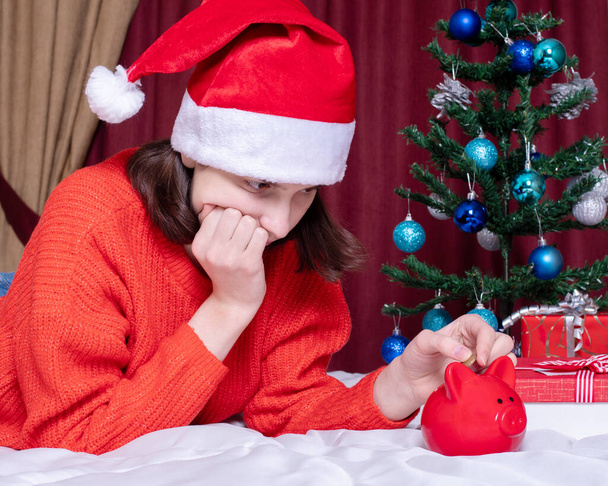 Saving money on Christmas gifts due to the pandemic crisis. Sad girl in a red christmas hat and sweater counts coins from a piggy bank in a home interior - Foto, immagini