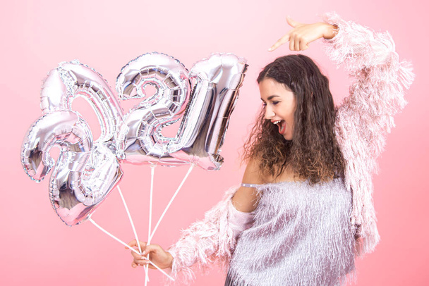 Beautiful cheerful young brunette with curly hair festively dressed on a pink background confidently posing with silver balloons in her hands from the numbers 2021 - Foto, afbeelding