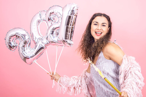 Hermosa chica morena alegre vestida festivamente con el pelo rizado en un fondo de estudio rosa posando con una vela de fuegos artificiales en la mano y bolas de plata de los números 2021, concepto de fiesta de año nuevo - Foto, Imagen