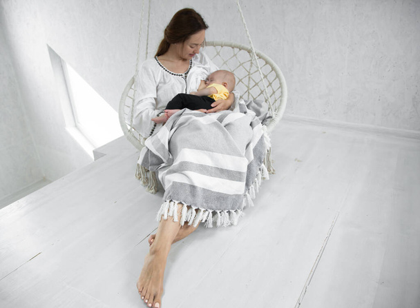 幸せなお母さんは白いハンモックで赤ん坊に日光の白い部屋の背景に毛布を与えます。幸せな母親の概念. - 写真・画像