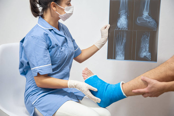 Ženský lékař v modrých lékařských šatech kontroluje zlomenou nohu a ukazuje mužskému pacientovi laterální projekci rentgen nohy a kotníku. - Fotografie, Obrázek