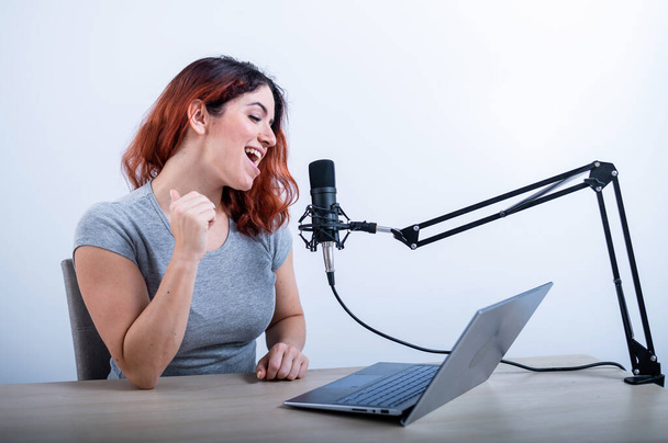 An emotional woman is broadcasting online on a laptop. Female radio presenter at the workplace - 写真・画像