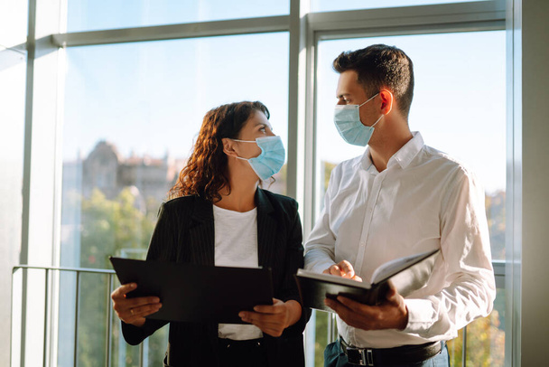 Two coworkers in protective face masks discussing project, sharing ideas, having pleasant conversation near elevator. Work in office during quarantine. Covid-19. - Photo, Image