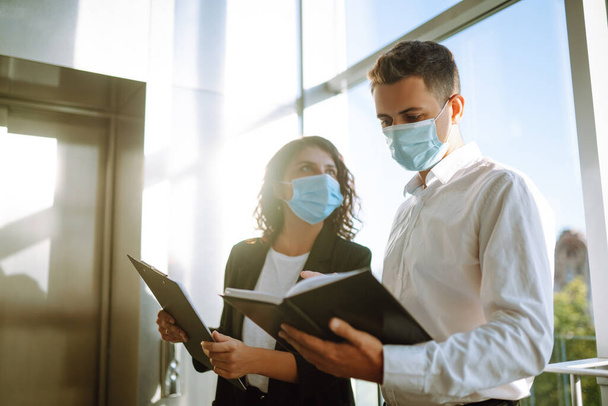 Two coworkers in protective face masks discussing project, sharing ideas, having pleasant conversation near elevator. Work in office during quarantine. Covid-19. - 写真・画像