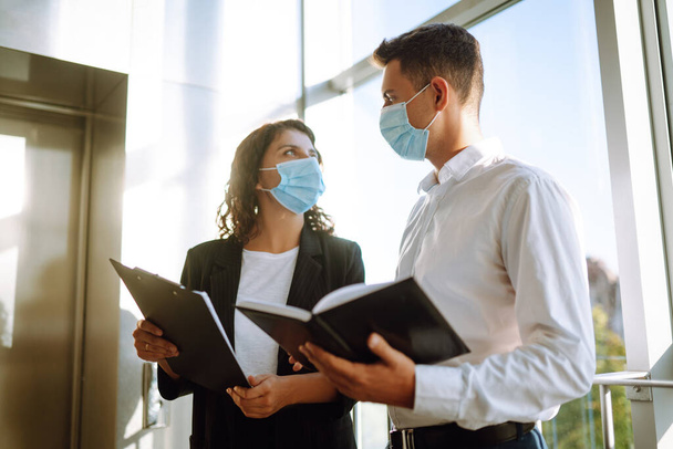 Two coworkers in protective face masks discussing project, sharing ideas, having pleasant conversation near elevator. Work in office during quarantine. Covid-19. - Фото, зображення