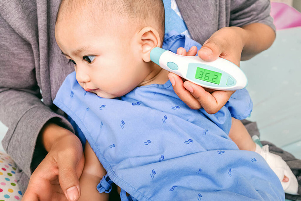Aasialainen poikavauva mittaa infrapunakuumetta hänen korvassaan. Keskity kasvoihin - Valokuva, kuva