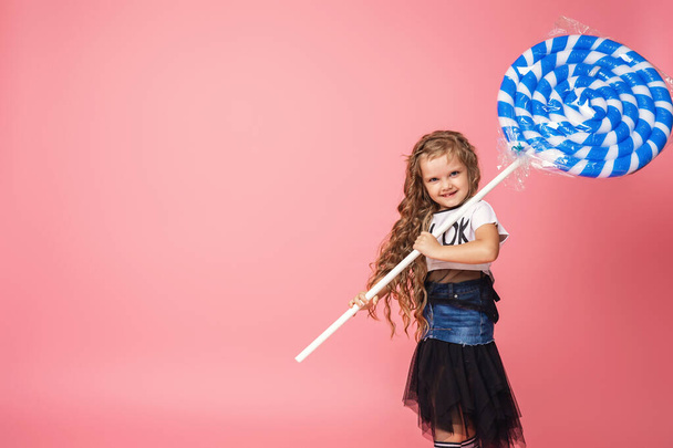 Stylish little girl 6 years old with curly hair and large pink Lollipop in fashionable clothes stands at full height on pink background. child smiles with toothless mouth. Emotional image. Copy space. - 写真・画像