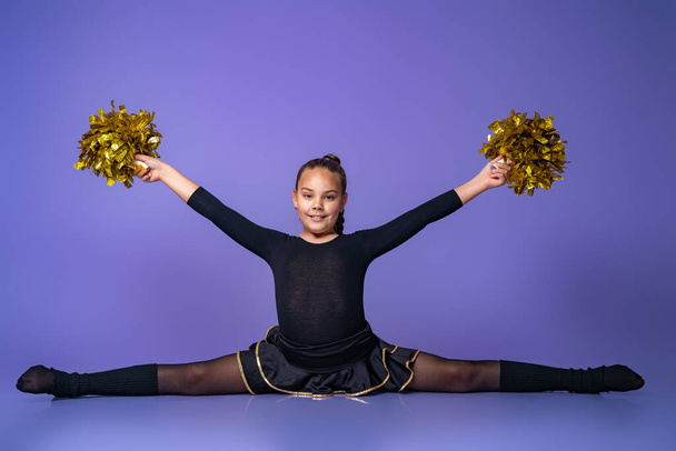 cheerleader teen girl sits on a twine in a Studio with gold POM-poms. a young gymnast does an exercise on a purple background. children's professional sports. Cheerleading - Photo, Image
