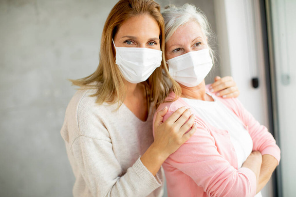 Starsza kobieta z opiekuńczą córką w domu w maskach medycznych jako ochrona przed koronawirusem - Zdjęcie, obraz