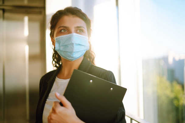 Young business woman wear face mask protect of coronavirus standing near elevator at modern office. Covid-19. - Zdjęcie, obraz