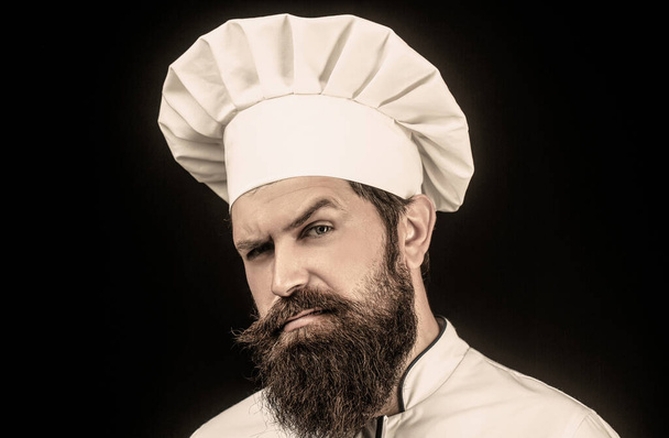 Kook hoed. Portret van een serieuze chef-kok. Baardkok, koks of bakker. Baard mannelijke chef-koks geïsoleerd op zwart. Vertrouwende bebaarde mannelijke chef-kok in wit uniform. Serieuze kok in wit uniform, chef-kok hoed - Foto, afbeelding