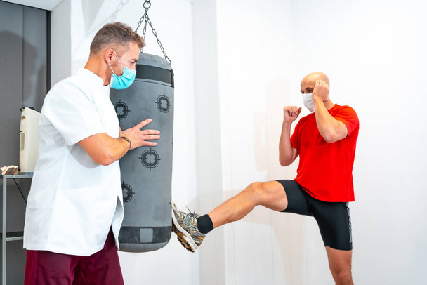 Physiotherapist with mask holding punching bag and kicking patient. Physiotherapy with protective measures for the Coronavirus pandemic, COVID-19. Osteopathy, sports quiromassage - Foto, immagini