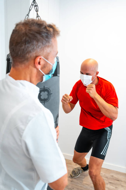 Physiotherapist with mask holding the punching bag for the patient to hit. Physiotherapy with protective measures for the Coronavirus pandemic, COVID-19. Osteopathy, sports quiromassage - Zdjęcie, obraz