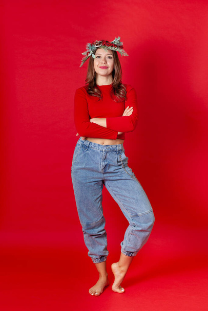full-length portrait of smiling young woman in Christmas wreath with her arms crossed on her chest, on a red background - Foto, Imagem