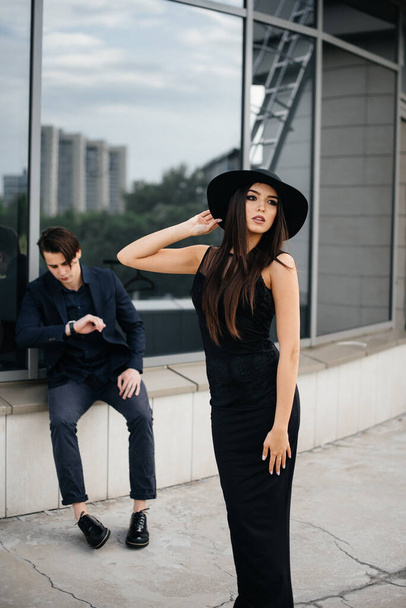 A beautiful, stylish pair of young people in black clothes and glasses stand against the background of an office building in the sunset. Fashion and style - Foto, Bild