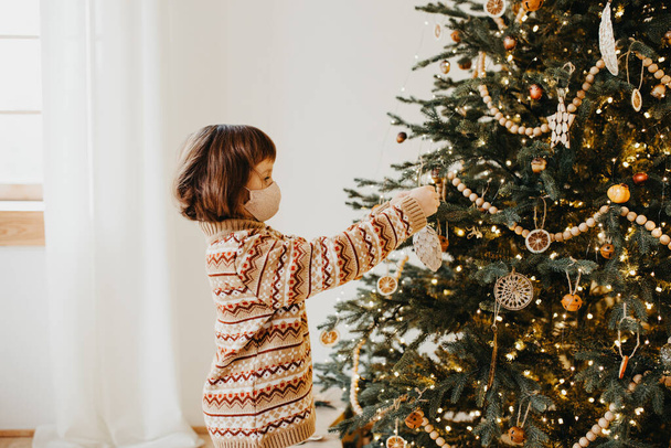 Child wearing warm sweater in protecting mask decorate christmas tree at home - Zdjęcie, obraz