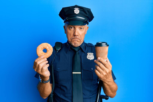Handsome middle age mature police man eating donut and drinking coffee depressed and worry for distress, crying angry and afraid. sad expression.  - Фото, изображение