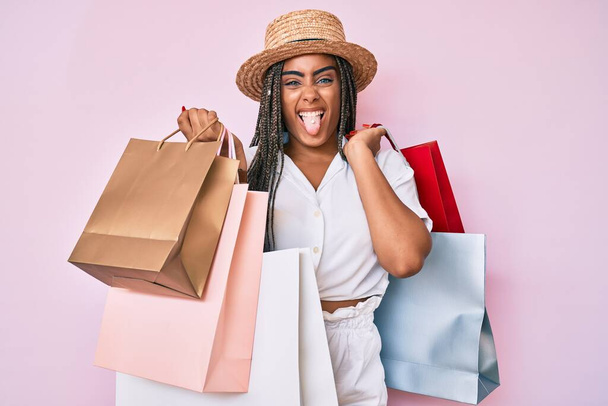 Junge afrikanisch-amerikanische Frau mit Zöpfen, die Einkaufstüten in der Hand halten, streckt die Zunge glücklich mit lustigem Ausdruck aus.  - Foto, Bild
