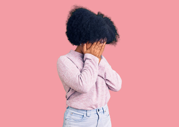 Νεαρή Αφροαμερικανή που φοράει καθημερινά ρούχα με λυπητερή έκφραση καλύπτοντας το πρόσωπο με τα χέρια ενώ κλαίει. έννοια της κατάθλιψης.  - Φωτογραφία, εικόνα
