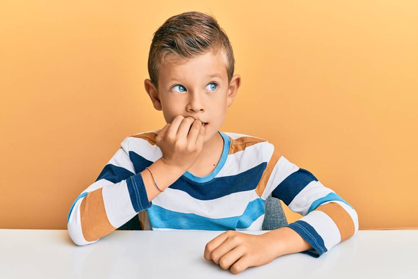 テーブルの上に座っているカジュアルな服を着ている愛らしい白人の子供は、口の中で爪をかむ手でストレスと緊張しています。不安の問題.  - 写真・画像