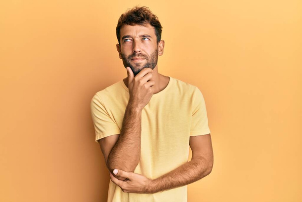 Handsome man with beard wearing casual yellow tshirt over yellow background with hand on chin thinking about question, pensive expression. smiling with thoughtful face. doubt concept.  - 写真・画像