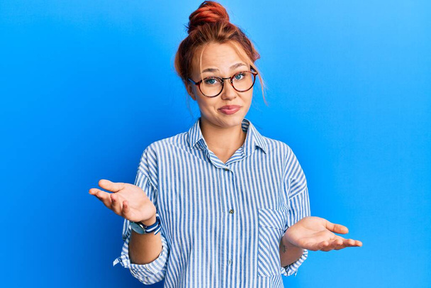 Young beautiful redhead woman wearing casual clothes and glasses over blue background clueless and confused expression with arms and hands raised. doubt concept.  - Photo, Image