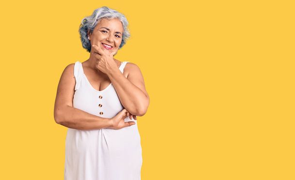 Starsza kobieta z siwymi włosami ubrana w luźne ubrania, wyglądająca pewnie przed kamerą, uśmiechnięta ze skrzyżowanymi ramionami i ręką podniesioną na podbródku. pozytywne myślenie.  - Zdjęcie, obraz
