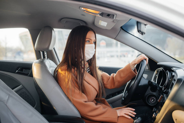 A young girl sits behind the wheel in the car in the mask during the global pandemic and coronavirus. Quarantine - 写真・画像