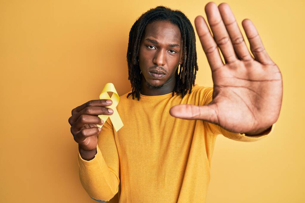 Hombre afroamericano con trenzas sosteniendo cinta amarilla de prevención suicida con mano abierta haciendo stop sign con expresión seria y confiada, gesto de defensa  - Foto, imagen