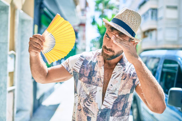 Молодий іспаномовний чоловік у відпустці за допомогою ручного вентилятора, що йде вулицею міста
 - Фото, зображення