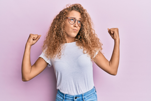 Menina adolescente branca bonita vestindo t-shirt branca sobre fundo rosa mostrando músculos braços sorrindo orgulhoso. conceito de aptidão.  - Foto, Imagem