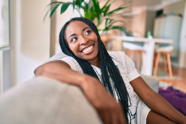 Joven afroamericana americana sonriendo feliz sentada en el sofá en casa - Foto, Imagen