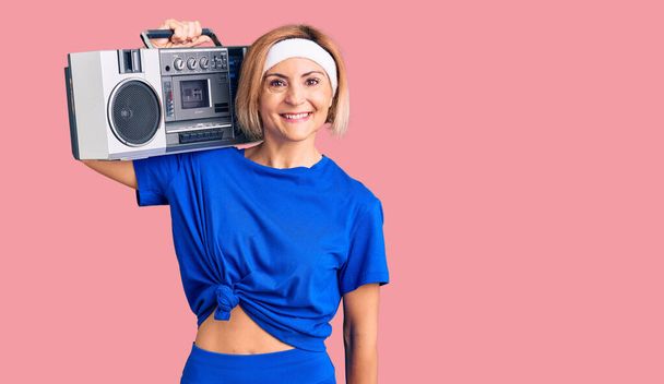 Young blonde woman wearing sportswear holding boombox, listening to music looking positive and happy standing and smiling with a confident smile showing teeth  - Foto, Imagem
