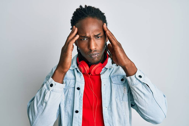 Afrykański Amerykanin z warkoczami słuchający muzyki za pomocą słuchawek cierpiących na ból głowy zdesperowany i zestresowany, ponieważ ból i migrena. ręce na głowę.  - Zdjęcie, obraz