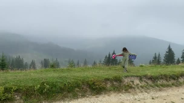 χαρούμενο κοριτσάκι τρέχει και πηδάει ανέμελα με ανοιχτές αγκάλες σε συννεφιασμένα βουνά. Κρατώντας σημαία των ΗΠΑ. επιλεκτική εστίαση. θόρυβος στις σκιές. - Πλάνα, βίντεο