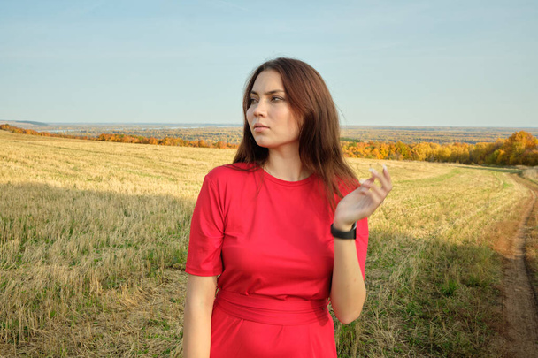 Young beautiful girl in a red dress stands in an autumn field - Фото, зображення