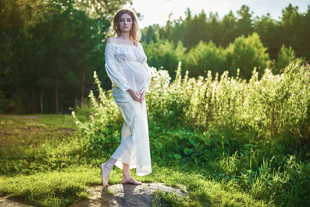 Young beautiful pregnant girl in a white dress and crown stands against the background of a waterfall  - Foto, Imagen